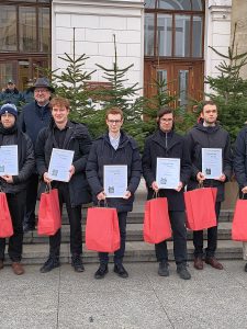 Stypendyści wraz z Rektorem PW stojący przed Gmachem Głównym PW na tle choinek trzymający dyplomy i upominki. 